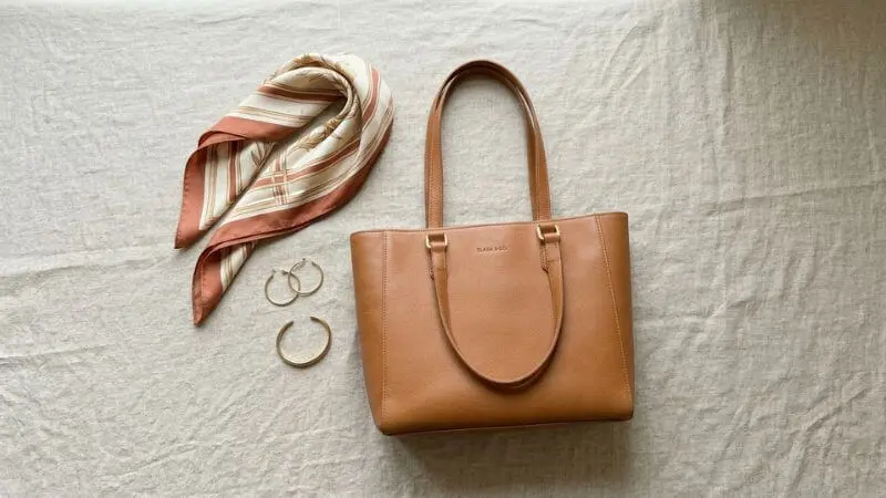 Flatlay de acessórios femininos sofisticados: bolsa tote caramelo, brincos dourados e lenço de seda em tons neutros