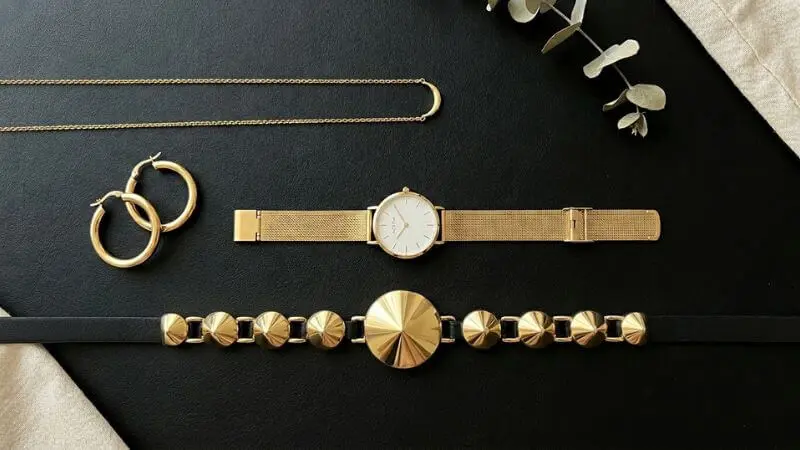 Flatlay de acessórios de metal polido dourado, incluindo brincos de argola e relógio, em estilo de luxo silencioso feminino minimalista.