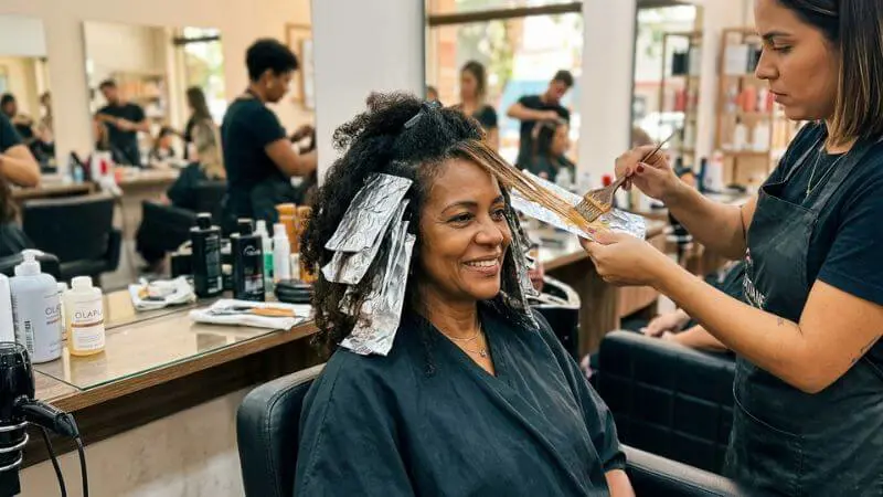 Cabeleireira aplicando mechas estratégicas próximas ao rosto de mulher madura em salão real
